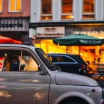 Coche clásico estacionado frente a una pizzería con luces cálidas al anochecer, con otros vehículos en la calle reflejando el ambiente urbano.