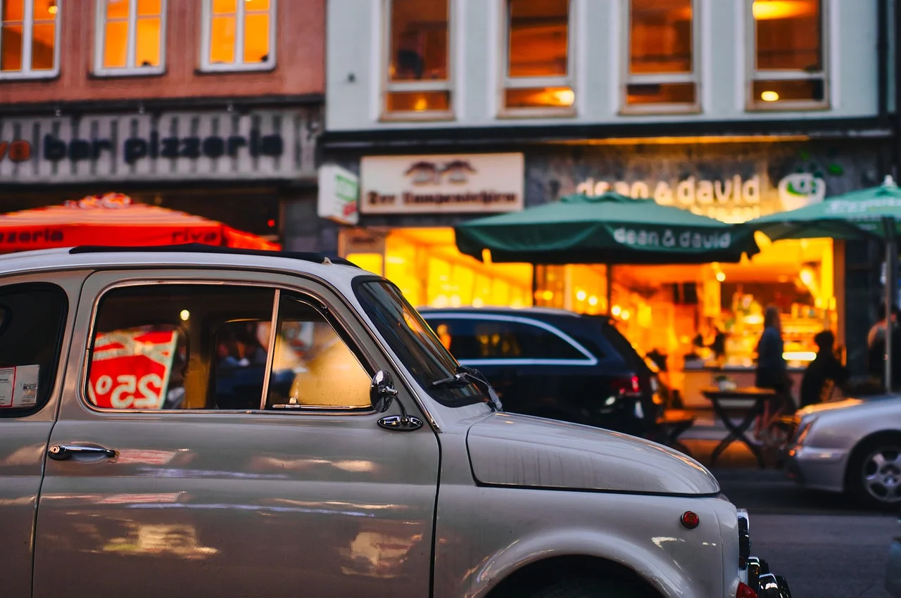 Coche clásico estacionado frente a una pizzería con luces cálidas al anochecer, con otros vehículos en la calle reflejando el ambiente urbano.