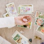 Tirada de cartas de tarot con ilustraciones florales y cristales energéticos sobre una manta, con una mano sosteniendo piedras de cuarzo.
