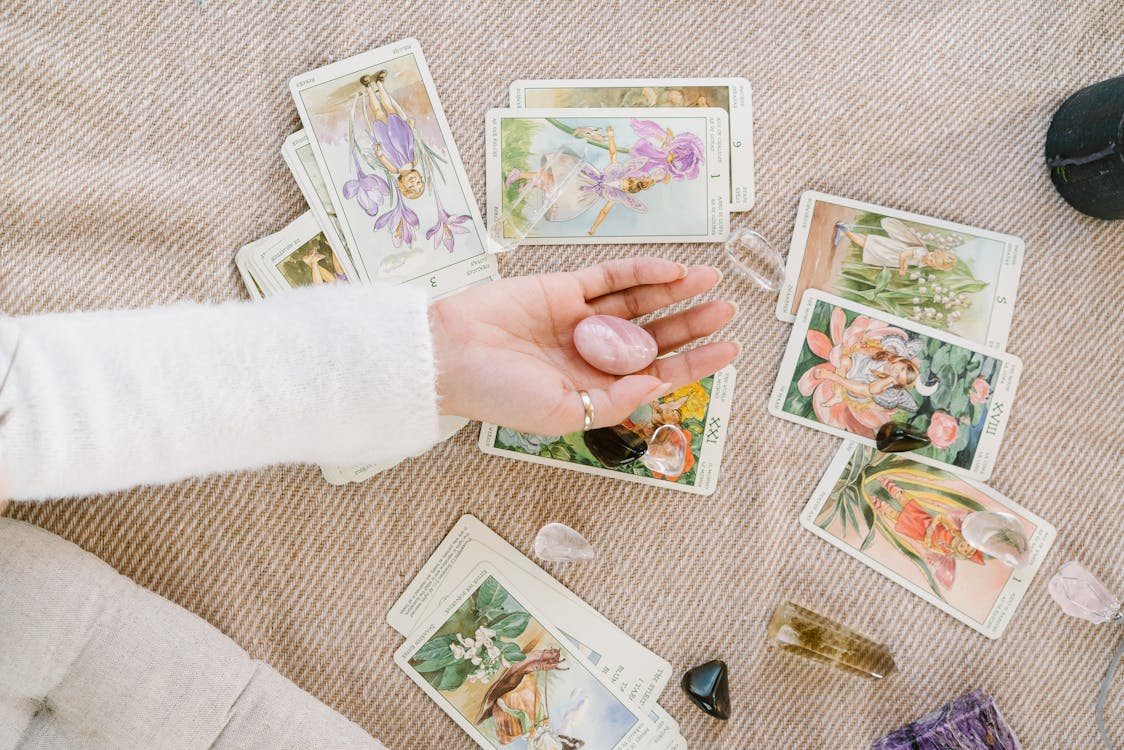 Tirada de cartas de tarot con ilustraciones florales y cristales energéticos sobre una manta, con una mano sosteniendo piedras de cuarzo.