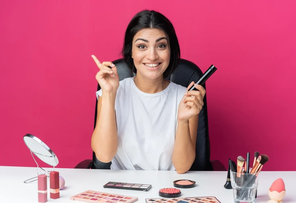 Mujer sonriente con productos de maquillaje en la mano, rodeada de brochas y paletas de colores, lista para aplicar su rutina de belleza.