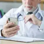 Médico usando un teléfono móvil mientras examina información relacionada con la salud, destacando la importancia de las aplicaciones de salud para profesionales médicos.