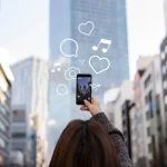 Persona tomando una foto con su teléfono móvil en una ciudad, rodeada de íconos de redes sociales flotando.